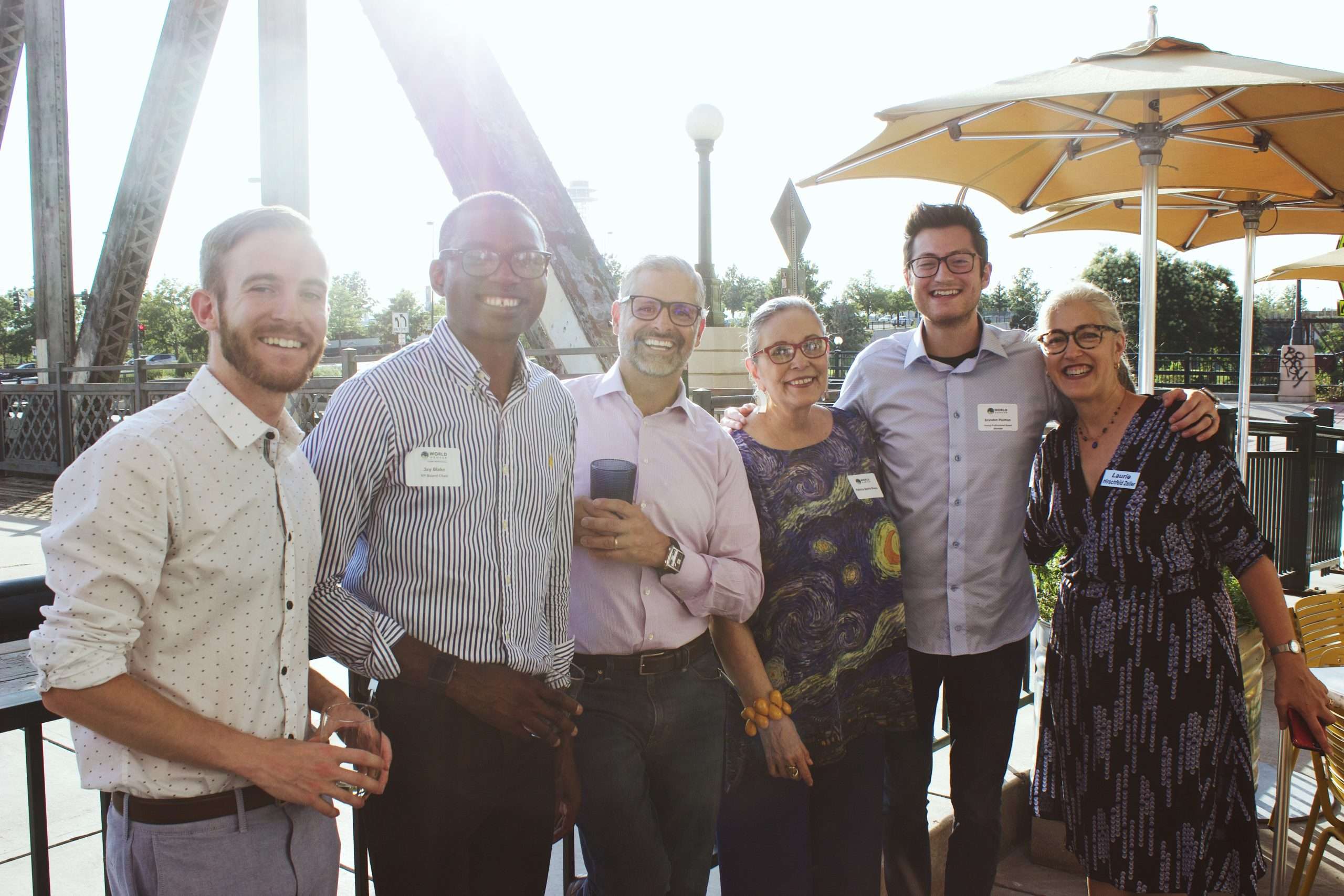 A group of 6 young professionals and WorldDenver members stand arm in arm smiling on a restaurant patio